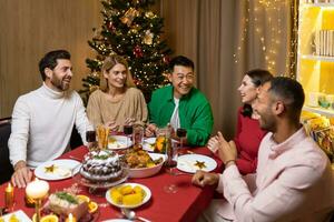 Happy young interracial people celebrating new year at home at festive table near Christmas tree. They hold glasses in their hands, laugh, communicate, rejoice photo