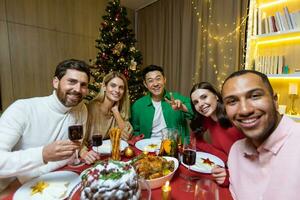 Festive selfie of a group of interracial people. Young men and women are sitting at the table in the apartment near the Christmas tree, holding glasses, looking at the camera, smiling. photo