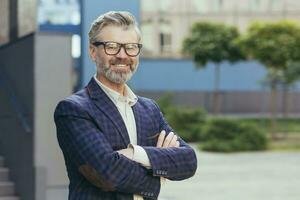 Portrait of mature grayhaired investor, senior man in glasses looking at camera and gesturing, businessman with crossed arms outside office building in business suit, successful boss banker. photo