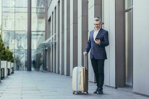 Business trip, senior grayhaired businessman outside hotel using phone, mature boss with big suitcase, investor in business suit and glasses. photo