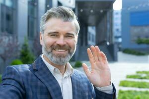 Video call of senior mature businessman, gray haired man looking phone camera and smiling, waving hand gesture of greeting, investor outside office building talking remotely with friends colleagues. photo