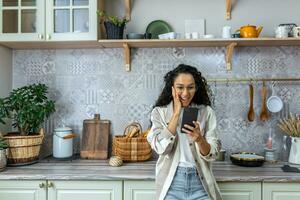A happy Latin American woman is standing in the kitchen at home, holding the phone in one hand, holding her head with the other hand. I am glad, I received an unexpected and joyful message, good news. photo