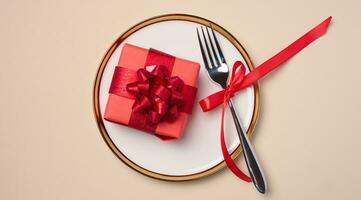 Red box with a gift on a round white plate and fork on a beige background, top view. photo