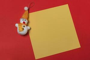 snowman and blank yellow sheet of paper on a red background, christmas concept photo