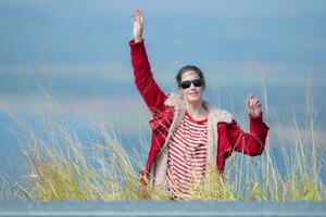 A cheerful young woman in a red jacket stands on top of a green hill, arms spread. photo