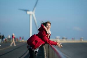 en el antecedentes de molinos de viento, un joven mujer en un rojo chaqueta es disfrutando su invierno vacaciones. foto