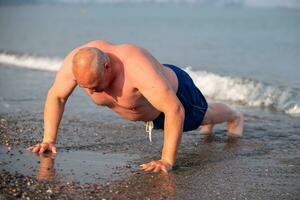 An elderly man of athletic build plays sports on the shore. Mature man doing push-ups outdoors. photo