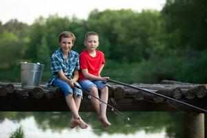dos Niños sentar con pescar varillas en un de madera puente y pescado en el lago. foto