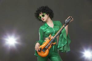 un estilo discoteca niña en un verde traje y un juguete guitarra posando en un gris antecedentes. foto
