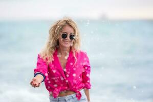 un hermosa mujer en Gafas de sol y un rosado camisa salpicaduras mar agua. foto