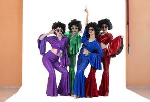 A group of girls in colorful flared suits and afro wigs pose in a photo studio. Disco style from the eighties or seventies.