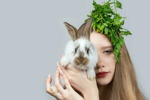 On a gray background, a close-up portrait of a rural girl with a bunch of parsley on her head and a small rabbit. photo