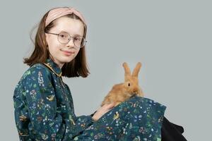 Beautiful little girl in glasses plays with a red rabbit on a gray background. photo