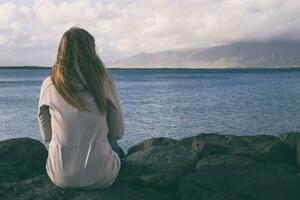 Woman sitting at the coastline in Reykjavik and looking at the sea.Image contains little noise because of high ISO set on camera and it is toned. photo