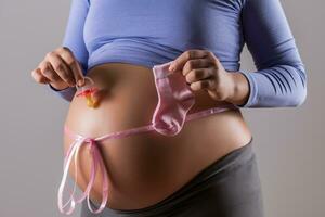 Image of stomach of pregnant woman with a pink ribbon holding pacifier and socks for baby girl on gray background. photo