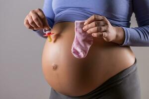 Image of stomach of pregnant woman with  holding pacifier and socks for baby girl on gray background. photo