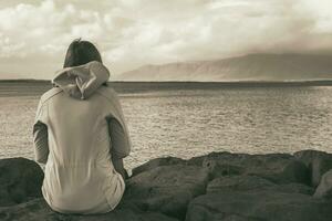 Sepia toned image of woman sitting at the coastline in Reykjavik and looking at the sea.Image contains little noise because of high ISO set on camera. photo