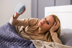 Angry woman in pajamas holding alarm clock in bed photo