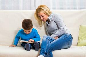 Happy mother and son are sitting on sofa and reading book togethe photo