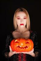 Beautiful girl dressed up like a vampire holding a pumpkin for halloween over black background. photo