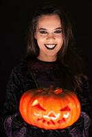 hermosa mujer vistiendo bruja atuendo para Víspera de Todos los Santos participación un escalofriante calabaza terminado negro antecedentes. foto