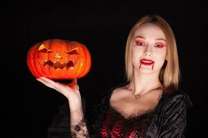 Portrait of beautiful woman dressed up like a vampire with bloody lips holding a pumpkin over black background. photo
