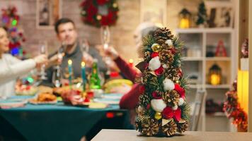 Little christmas tree on focus with family celebrating christmas in the background. Traditional festive christmas dinner in multigenerational family. Enjoying xmas meal feast in decorated room. Big family reunion photo