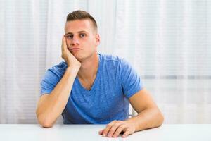 Depressed young man is sitting at the table and thinking about something. photo