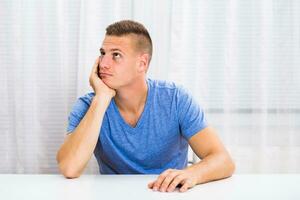 Depressed young man is sitting at the table and thinking about something. photo