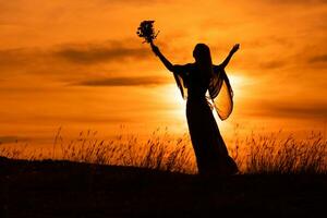 silueta de un mujer con brazos extendido participación flores y mirando a hermosa puesta de sol. foto