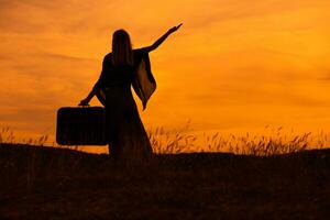 silueta de un mujer participación maleta con brazo elevado y mirando a hermosa puesta de sol. foto
