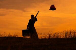 Silhouette of a woman holding suitcase and  heart shaped balloon while looking at beautiful sunset. photo