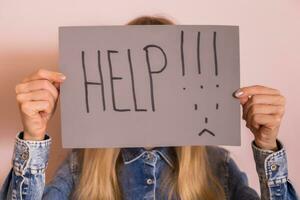 Woman holding paper with word help and sad face while standing in front of the wall. photo