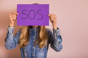 Woman holding paper with word SOS while standing in front of the wall. photo