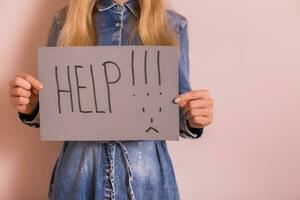 Woman holding paper with word help and sad face while standing in front of the wall. photo