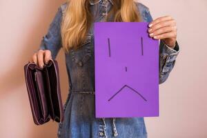 Woman holding empty wallet and sad face on paper while standing in front of wall. photo