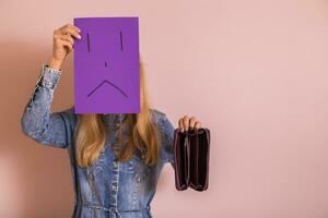 Woman holding empty wallet and sad face on paper while standing in front of wall. photo
