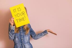 Woman holding paper with text it is your time while standing in front of the wall. photo