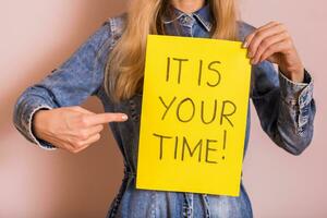 Woman holding paper with text it is your time while standing in front of the wall. photo