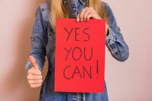 Woman holding paper with text yes you can and showing thumb up while standing in front of the wall. photo