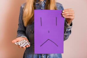 Woman holding pills and sad face on paper while standing in front of wall. photo