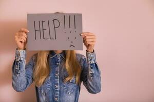 Woman holding paper with word help and sad face while standing in front of the wall. photo