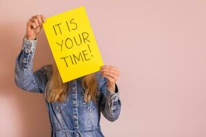 Woman holding paper with text it is your time while standing in front of the wall. photo