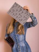 Woman holding empty wallet and paper with question marks while standing in front of wall. photo