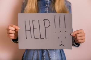 Woman holding paper with word help and sad face while standing in front of the wall. photo