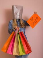 juguetón mujer participación compras pantalones con bolso en su cabeza y papel con texto grande rebaja mientras en pie en frente de el pared. foto