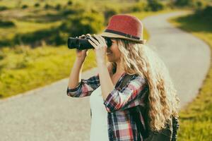 Beautiful young woman at the country road enjoys looking through binoculars. photo
