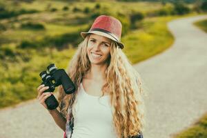 Beautiful young woman at the country road enjoys looking through binoculars.Toned image. photo