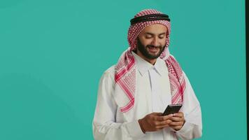 arabicum person i traditionell utrusta innehav smartphone i händer och skriver i uppkopplad social media chatt. muslim vuxen stående med mobil telefon och textning på webbsida, internet webbplats. video