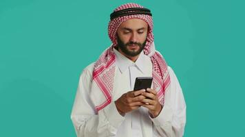 arabisch Kerl halten seine Handy, Mobiltelefon Gerät, Surfen über Sozial Medien Plattformen und chatten online. Muslim mit üblich bekleidung Verwendet Smartphone zu Zugriff global Netz, Gefühl aufgeregt auf Kamera. video